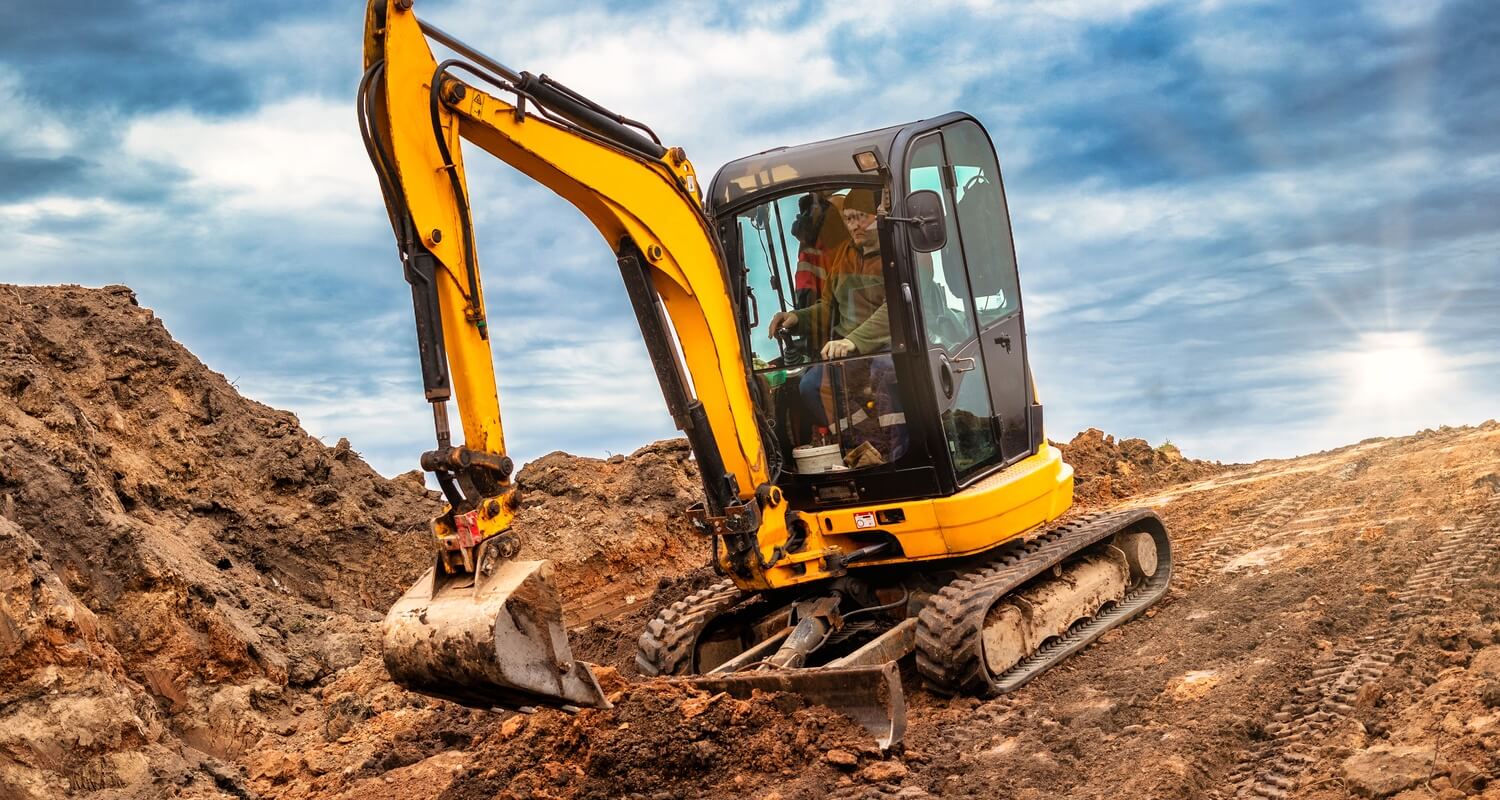 - Machinery Asia A person operating a yellow mini excavator on a sloped dirt mound set against a blue sky with clouds and bright sun.